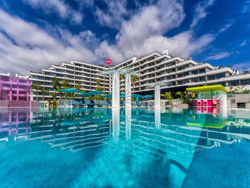 Moderní hotel s velkým bazénem a modrou oblohou v pozadí.