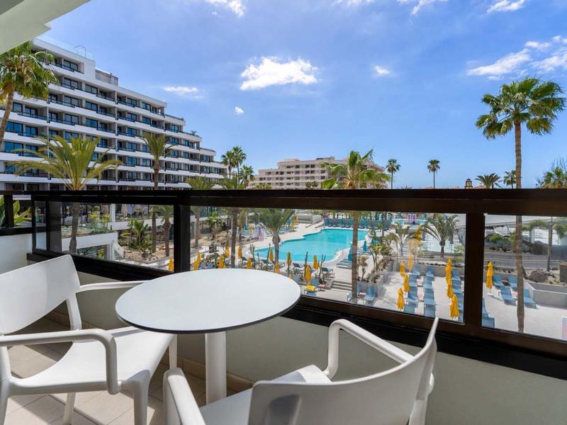 Balkon s bílým stolem a židlemi, výhled na bazén a palmy pod modrou oblohou.