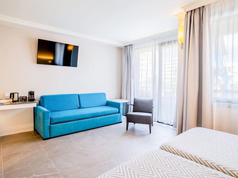 Světle hotelový pokoj s modrou pohovkou, křeslem, televizí na zdi a velkými okny s záclonami.