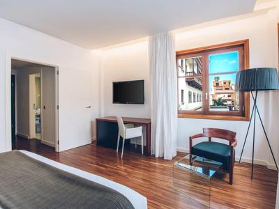 Helles Hotelzimmer mit Holzboden, Flachbildschirm, Fenster, Vorhängen, Stuhl und Stehlampe.
