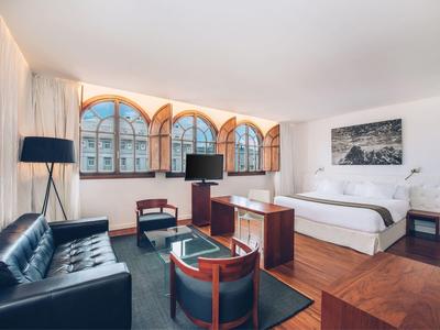 Modern eingerichtetes Hotelzimmer mit großen Rundbogenfenstern, Bett, Sofa und Sitzbereich.