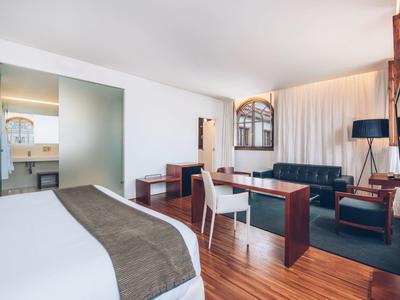Hell eingerichtetes Hotelzimmer mit Holzboden, blickdichtem Vorhang, Sitzgruppe und großem Bett.