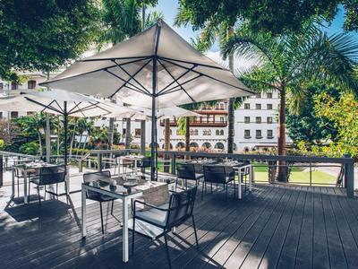 Freiluft-Café mit weißen Sonnenschirmen und schwarzen Stühlen auf Holzboden unter Bäumen.