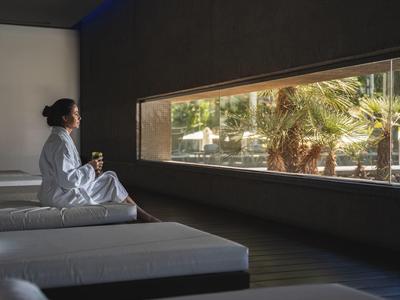 Frau in Bademantel sitzt auf Liege in Spa, schaut durch langes Fenster auf Palmen und Gebäude.