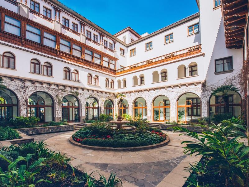Patio interior de hotel con arcos, jardín central y arquitectura tradicional bajo cielo azul.