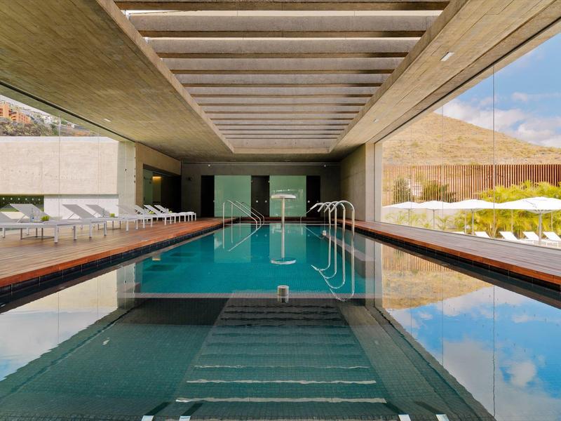 Piscina interior moderna con tumbonas y grandes ventanas que ofrecen vista al exterior.