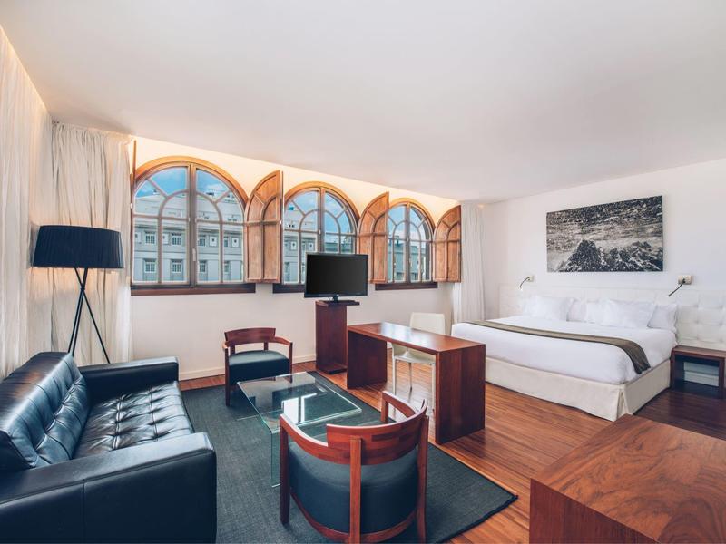 Habitat luminoso con cama grande, sofá de cuero, sillones y ventanas grandes con vista a la ciudad.