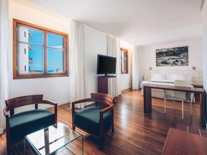 Habitación de hotel luminosa con suelo de madera, dos sillones, mesa de vidrio y cama con vista a la ciudad.