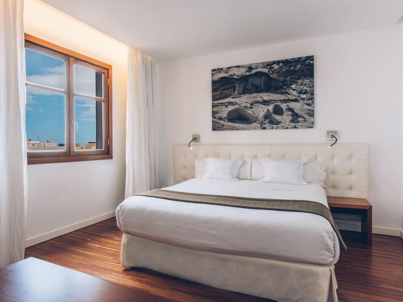 Habitación de hotel luminosa con cama doble, suelo de madera y vista al mar por la ventana.