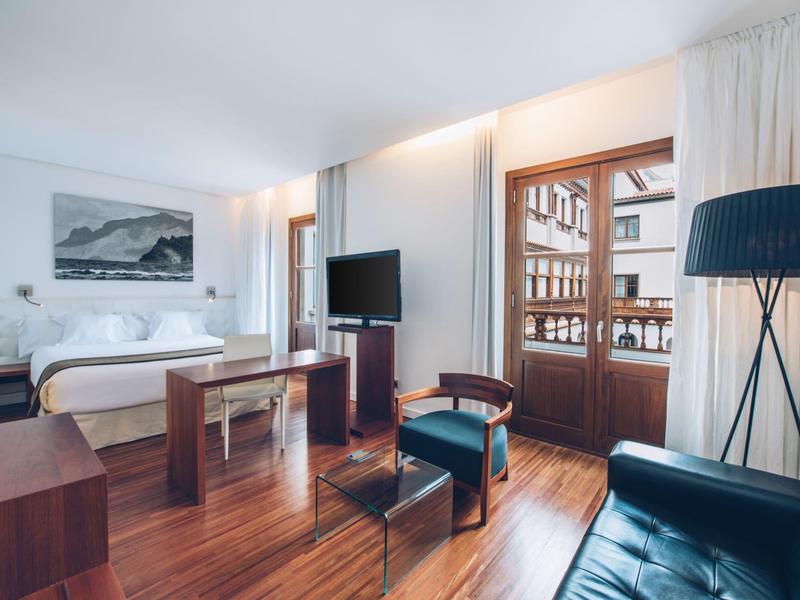 Habitación de hotel luminosa con suelo de madera, cama doble, sofá, televisor y balcón con vista a edificios.