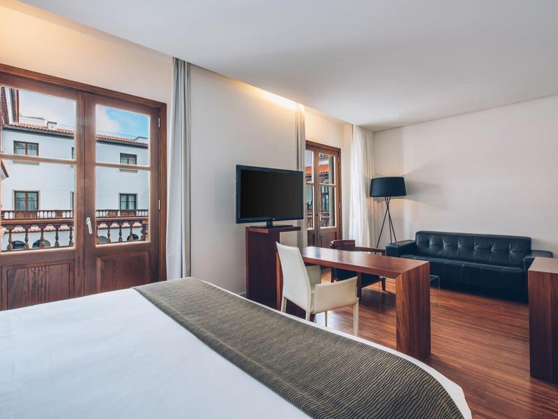 Habitación de hotel moderna con cama, sofá, escritorio y ventana grande con vista a la ciudad.