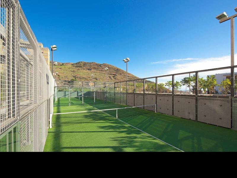 Cancha de tenis al aire libre con suelo verde, rodeada de cercas y montañas bajo cielo azul.