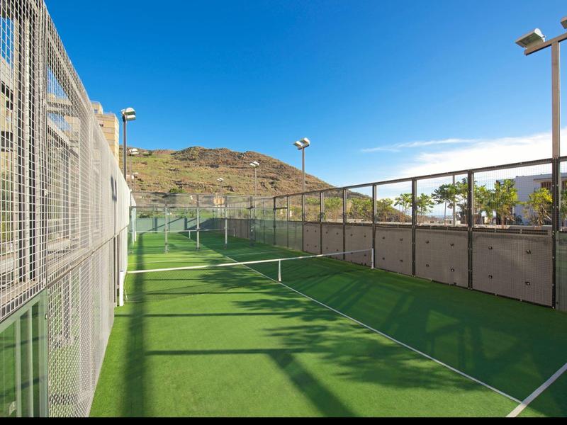 Una cancha deportiva verde al aire libre con cercas y vista a colinas bajo un cielo despejado.