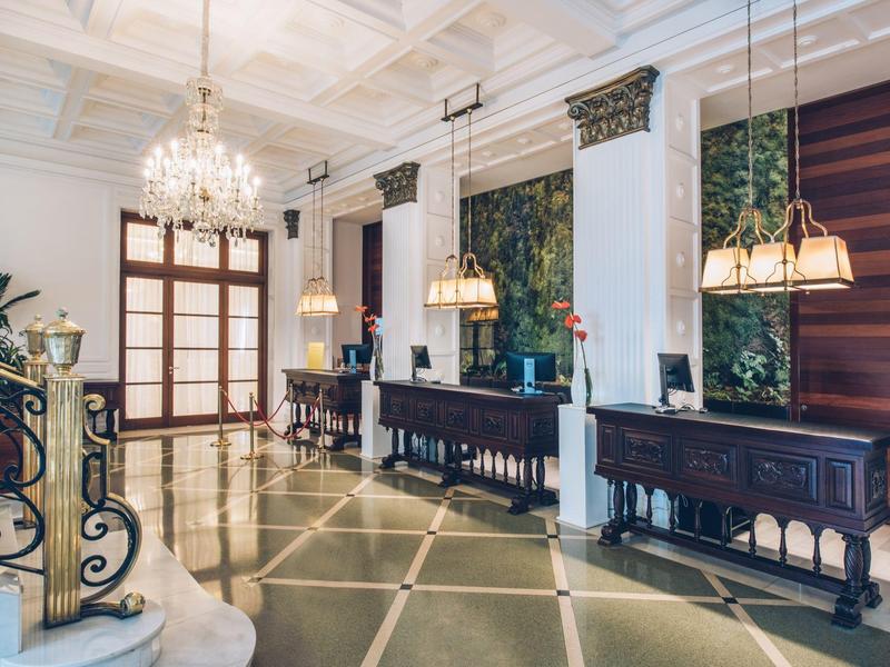 Elegante recepción de hotel con candelabros, techos altos y muebles de madera oscura en estilo clásico.