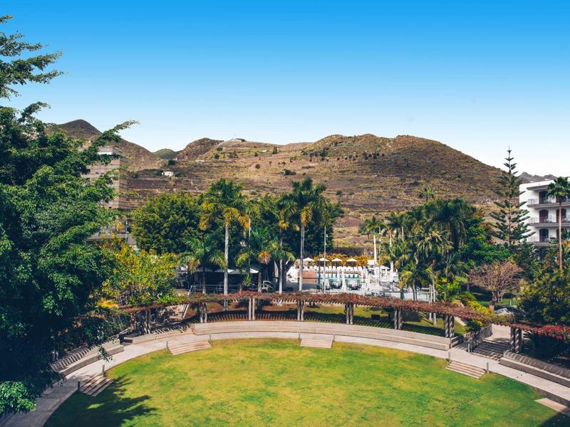 Área de parque verde con bancos, palmeras y montañas al fondo bajo cielo despejado.