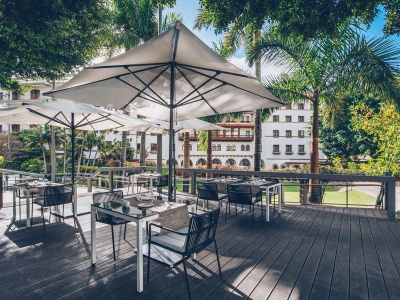 Terraza soleada de hotel con mesas, sillas y grandes sombrillas en un área verde.