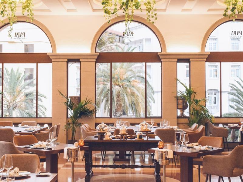 Restaurante elegante con grandes ventanas, vista a palmeras y mesas elegantemente preparadas.