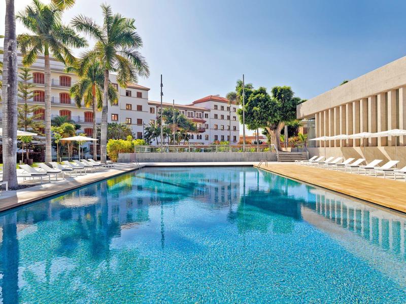 Piscina al aire libre con palmeras, rodeada de tumbonas y edificios vacacionales