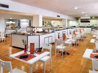 Großes, modernes Restaurant mit weißen Tischen, Stühlen und orangenen Tischsets, hell und sauber.