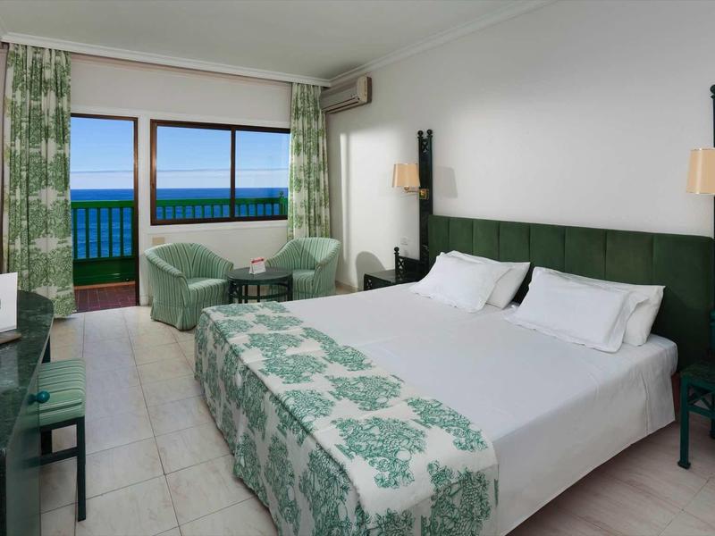 Hotelzimmer mit großem Bett, grün gemusterten Vorhängen, zwei Sesseln und Meerblick durch Fenster.