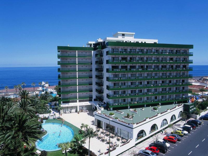 Großes Hotelgebäude mit Balkonen, Pool, Palmen und Meer im Hintergrund bei klarem Himmel.
