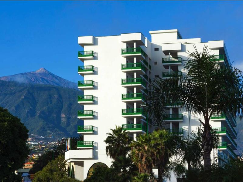 Hohes weißes Gebäude mit grünen Balkonen, umgeben von Palmen und Bergen im Hintergrund.