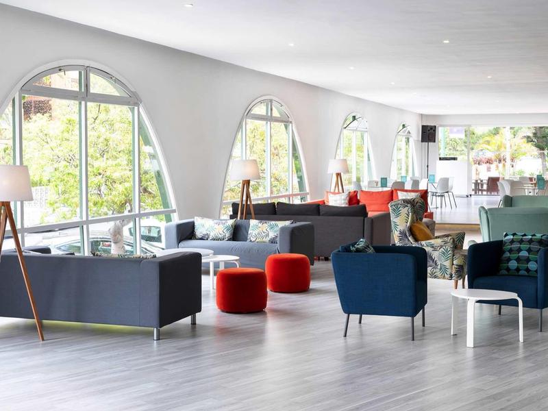 Heller Loungebereich mit großen Rundbogenfenstern, bunten Sesseln, Sofas und Holzvertäfelung.