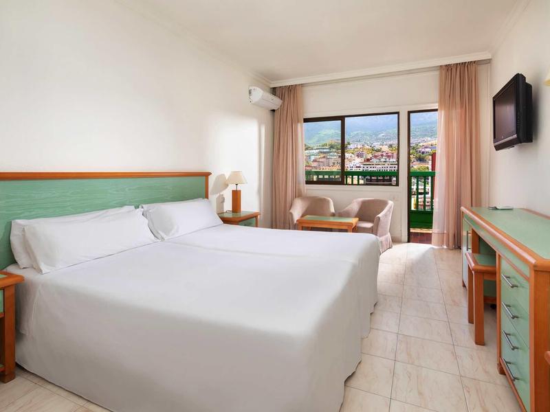 Helles Hotelzimmer mit Doppelbett, Balkon, Fernseher, Schreibtisch und Sitzbereich mit Blick auf die Stadt.