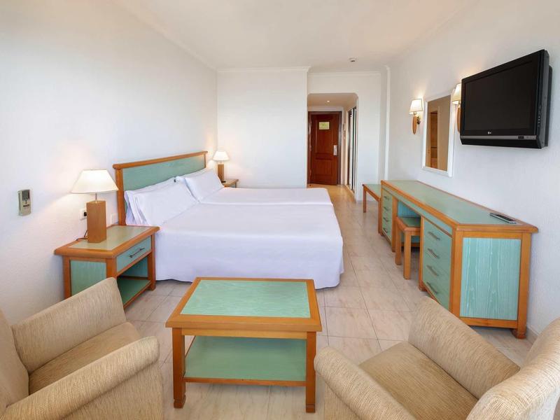 Hell eingerichtetes Hotelzimmer mit zwei Einzelbetten, Stühlen, Tisch und Wandfernseher.