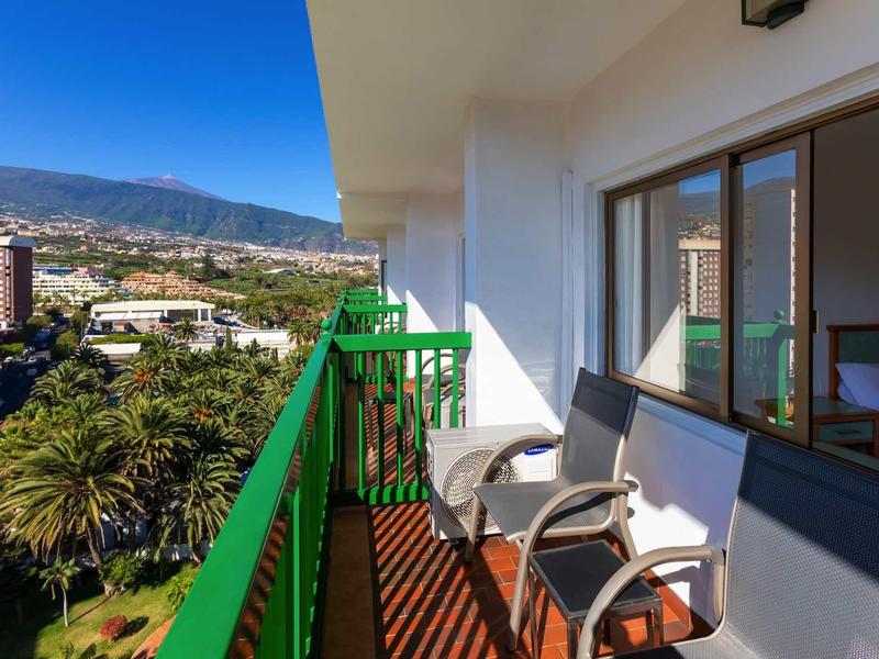 Balkon mit grüner Brüstung, zwei Stühlen und Blick auf Stadt, Palmen und Berge unter blauem Himmel.