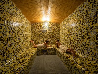 Dos personas descansan en una sauna con mosaicos amarillos y azules y techo de madera.