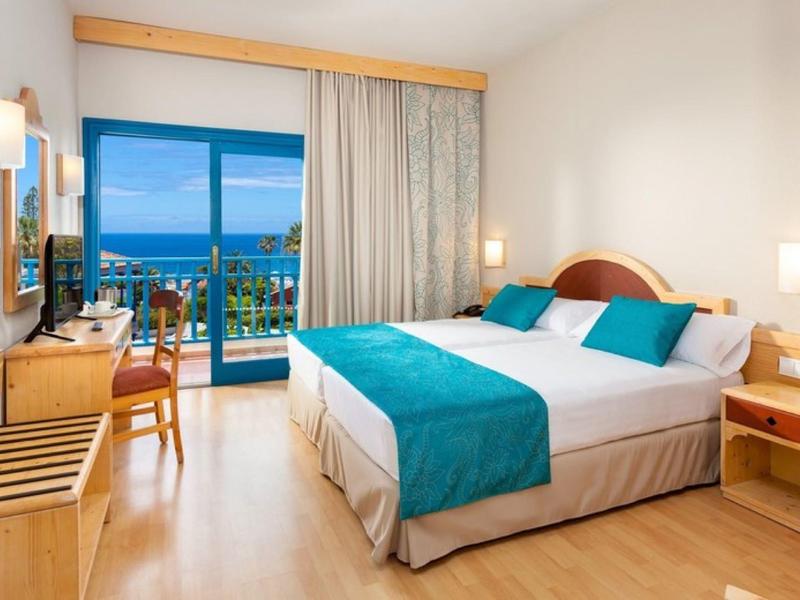 Habitación de hotel luminosa con cama doble, balcón con vista al mar y decoración azul y blanca.