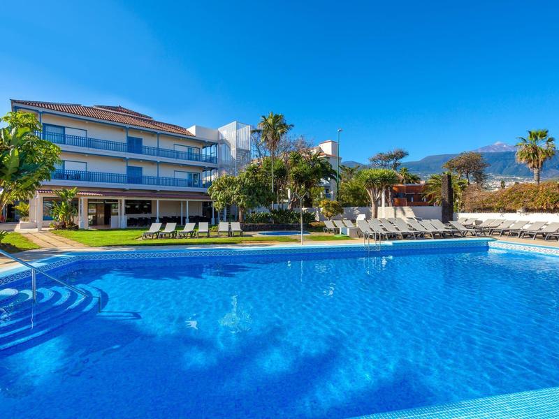 Complejo hotelero moderno con gran piscina rodeada de palmeras y jardín soleado.