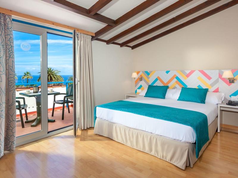 Habitación de hotel luminosa con cama doble, balcón con vista al mar y decoración colorida.