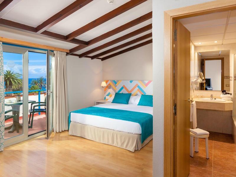 Habitación de hotel luminosa con cama doble, suelo de madera y balcón con vista al mar.