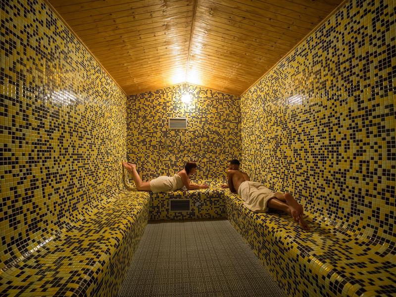 Dos personas descansan en una sauna con mosaicos amarillos y azules y techo de madera.