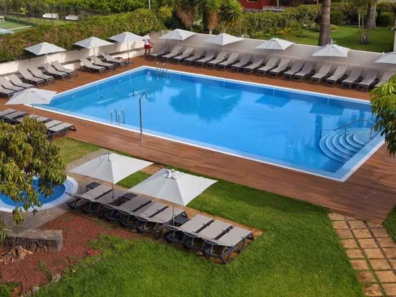 Piscina rectangular moderna con agua azul, rodeada de tumbonas en terraza de madera y césped.