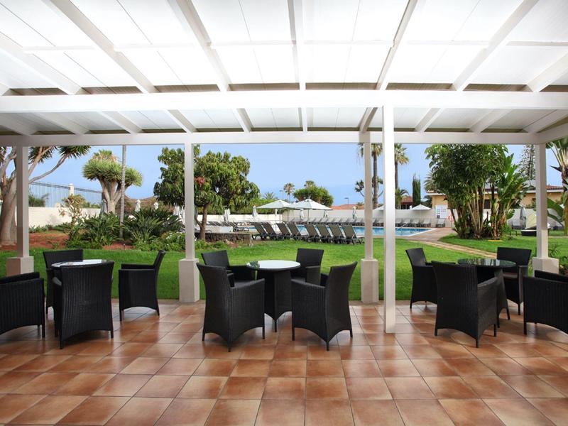 Terraza cubierta con áreas de asientos y vista a un jardín y piscina de un hotel.