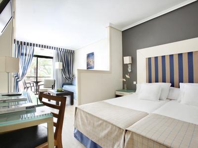 Chambre d'hôtel lumineuse avec lit, bureau, chaises et grande fenêtre avec rideaux bleus.