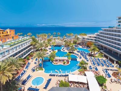 Großer Hotelpool mit Sonnenschirmen, Liegen, Palmen und Meer im Hintergrund bei klarem Himmel.