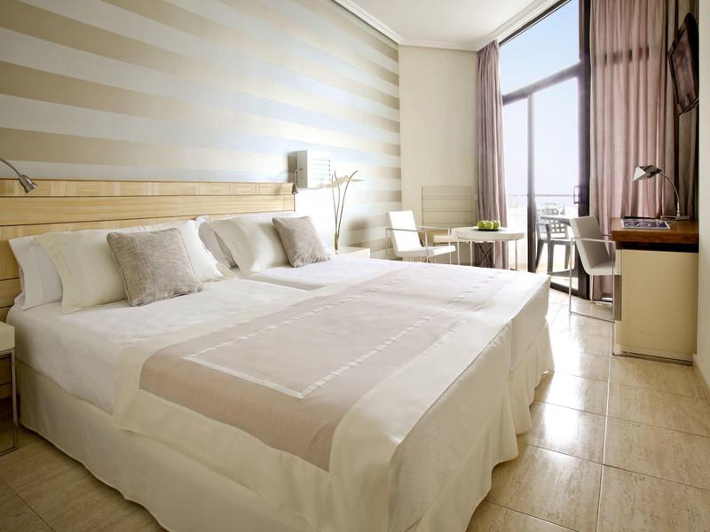 Chambre d'hôtel lumineuse avec grand lit, mur rayé et fenêtre avec chaises à côté.