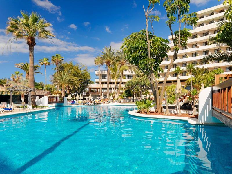 Piscine extérieure d'hôtel entourée de palmiers, chaises longues et bâtiments à plusieurs étages sous ciel bleu.