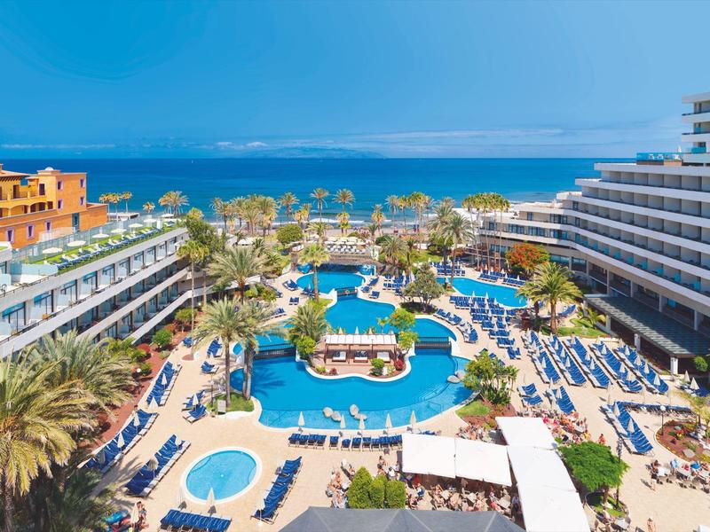 Grand espace piscine d'hôtel avec transats, palmiers et vue sur l'océan en arrière-plan.