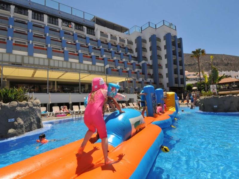 Kinder spielen auf buntem aufblasbarem Parcours im blauen Pool vor Hotelgebäude bei sonnigem Wetter.