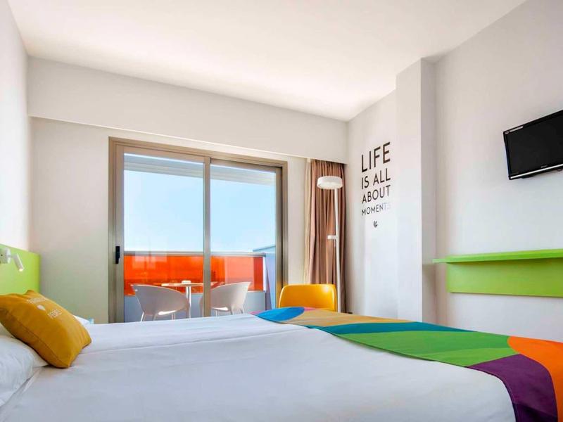 Helles, modernes Hotelzimmer mit Doppelbett, bunten Kissen, Balkon und Meerblick.