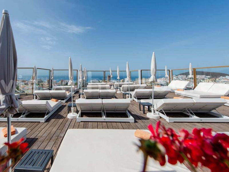 Moderne Dachterrasse mit weißen Liegen, Holzterrasse, Sonnenschirmen und blauem Himmel im Hintergrund.