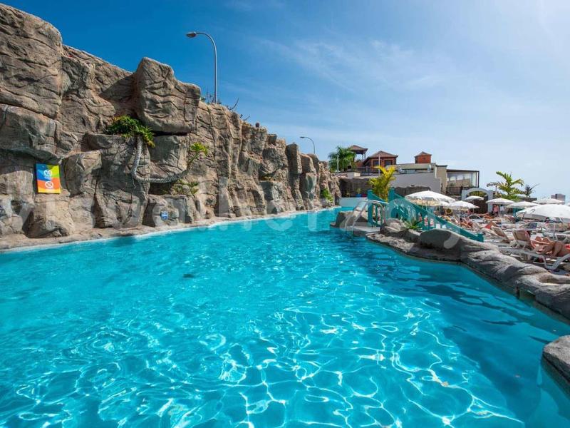 Klarer Pool mit Felsenwänden, Liegestühlen und blauem Himmel im sonnigen Außenbereich.