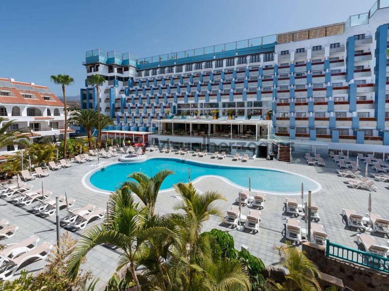 Großes Hotel mit blauem Außenpool, Liegestühlen und Palmen unter klarem Himmel.