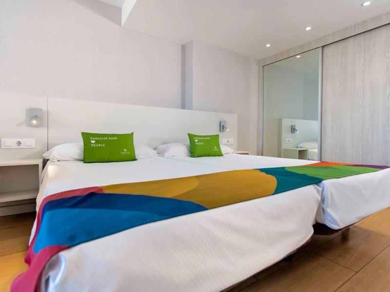 Helles Schlafzimmer mit Doppelbett, bunten Decken und weißen Wänden, Schiebetür und Holzboden.