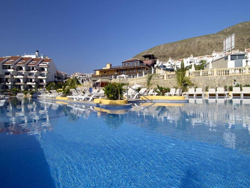 Großer blauer Pool vor weißen Hotels und Hügeln unter klarem, blauem Himmel.